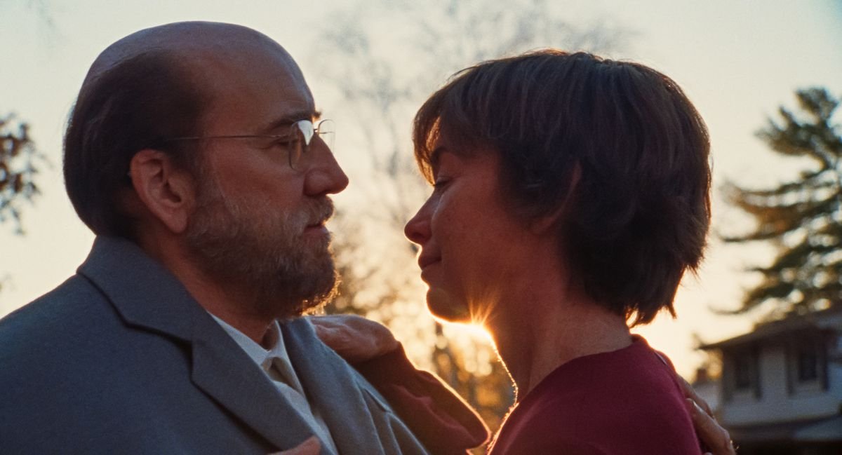 Balding professor Paul Matthews (Nicolas Cage) and his wife Janet (Julianne Nicholson) lean toward each other in close-up with the sun providing a highlight behind them in A24’s movie Dream Scenario