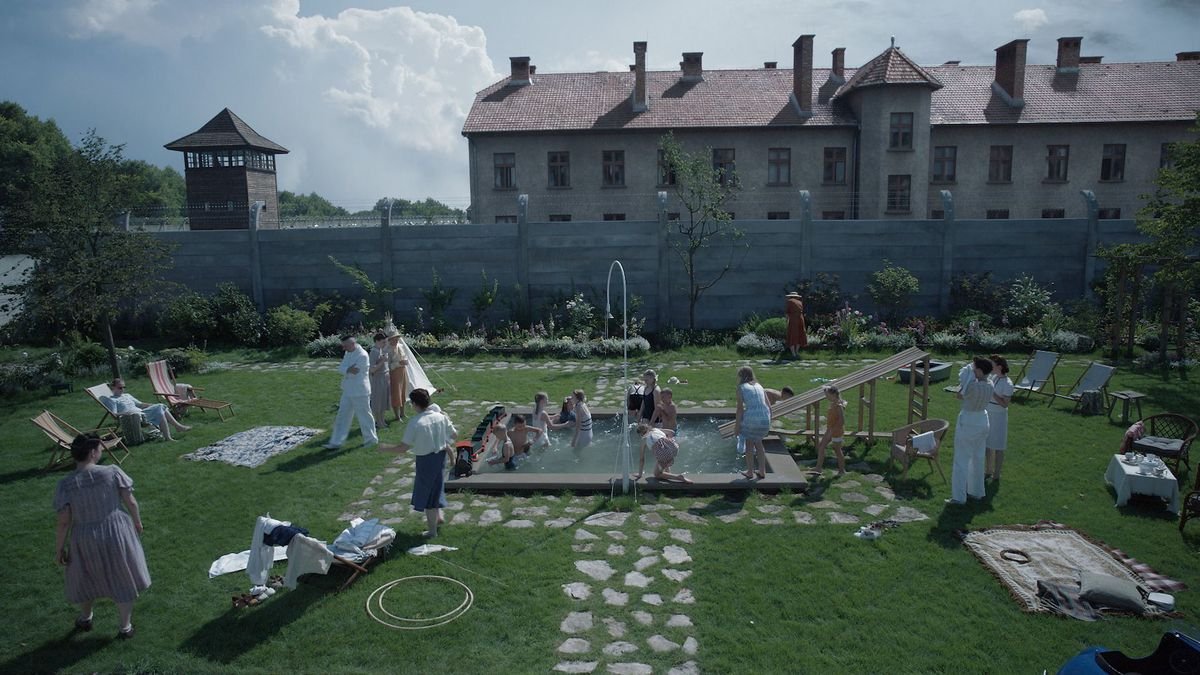 Several people stand in a walled garden with the towers of Auschwitz behind them in The Zone of Interest