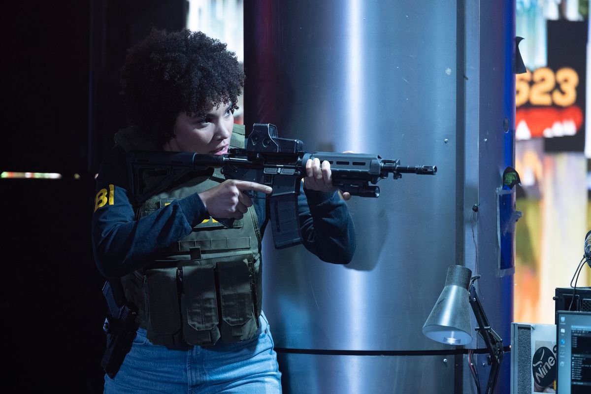 FBI Agent Verona Parker (Emmy Raver-Lampman), wearing a bulletproof vest and an FBI uniform, aims an assault rifle in The Beekeeper.