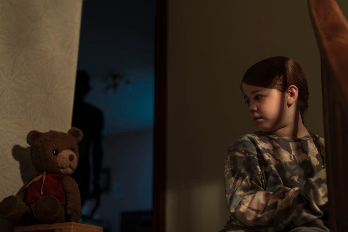 A young girl played by Pyper Braun sits at the top of the stairs next to a teddy bear while an ominous shadowy figure lurk behind her in Imaginary