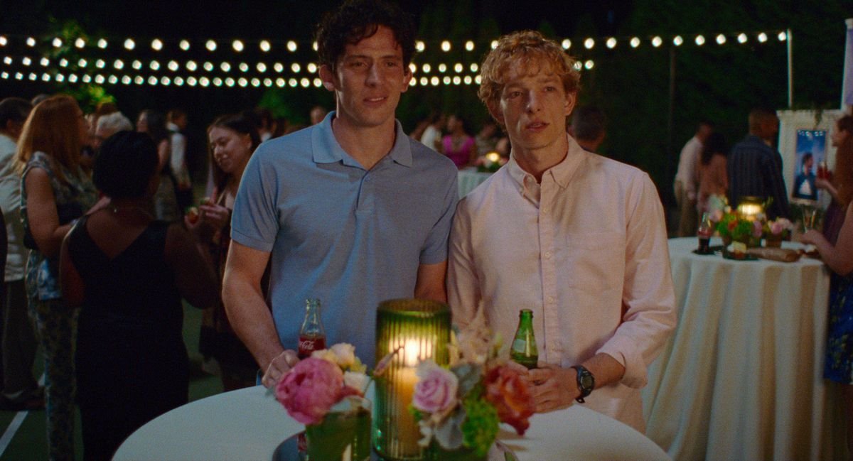 Tennis partners Art (Mike Faist) and Patrick (Josh O’Connor) sit at an outdoor table at a party with a candle in front of them and strings of lights behind them, staring admiringly at something (OK, it’s Zendaya) offscreen in Challengers