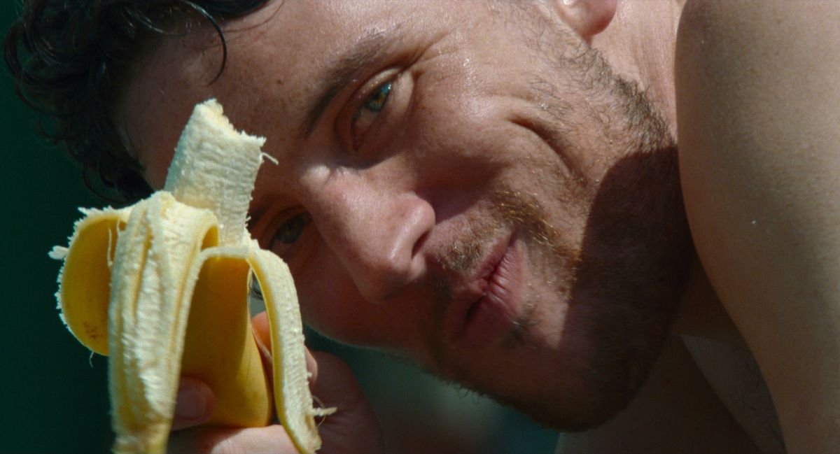 Patrick (Josh O’Connor), shirtless and scruffy and smiling, Very Pointedly eats a banana in extreme close-up in a shot from Challengers