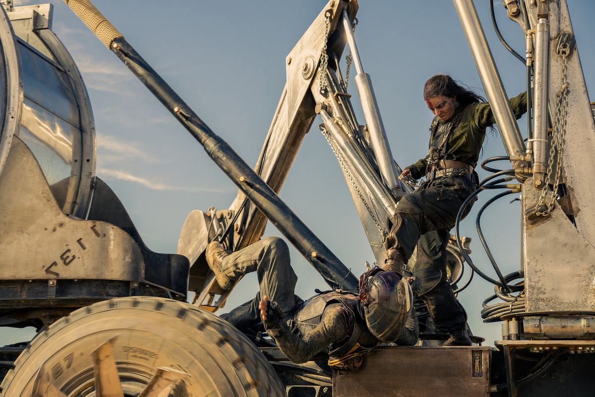 Furiosa (Anya Taylor-Joy), a leather-clad warrior with long braided hair, stands on the back of a steam shovel, kicking a helmeted, prone adversary in the face, in George Miller’s Furiosa