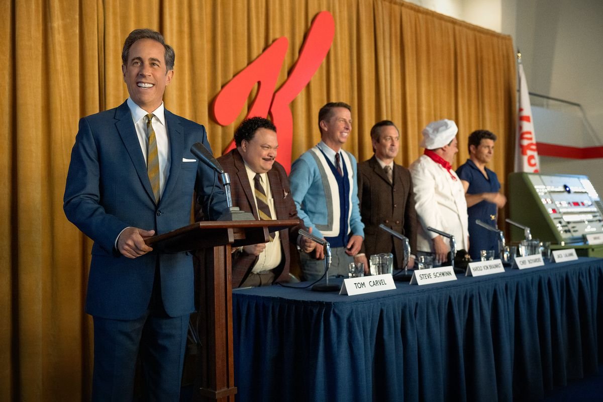 Seinfeld stands behind a podium in front of the Kellogg’s K logo alongside a panel of other people in the Netflix movie Unfrosted.