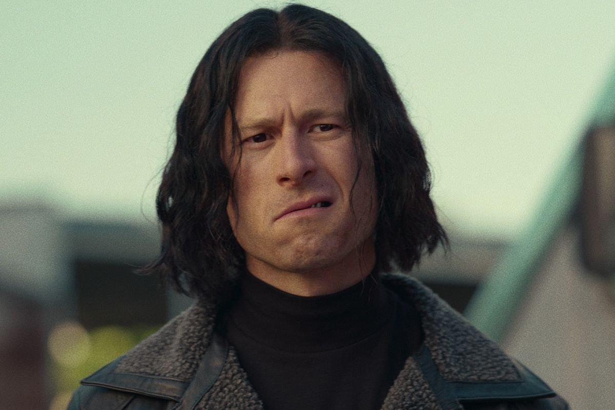 Glen Powell in a long black wig, leather coat, and weird sneer in Hit Man