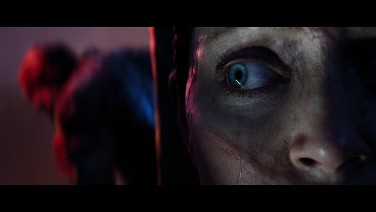 A zoomed-in shot of part of Senua’s face, her right eye shifted in panic to her right side. Behind her, there is a blurry figure