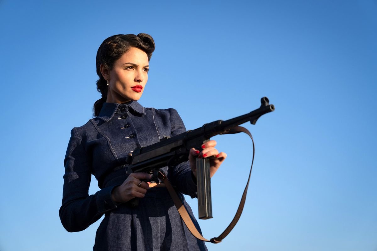 Eiza González, in a smart 1940s dress and scarlet lipstick, holds a machine gun in The Ministry of Ungentlemanly Warfare