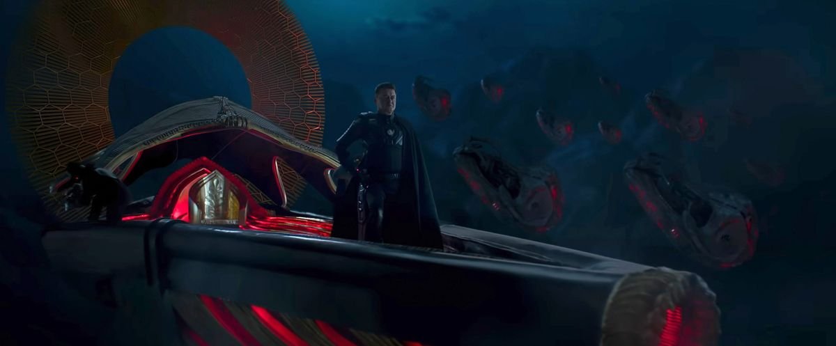 In a promo image for the Indian sci-fi blockbuster Kalki 2989 AD, a man in black clothing and a long black cape stands in a dark, V-shaped object that looks like a single-person spaceship with a crimson-lined interior and closing crimson highlights. Behind him in the darkness, a group of similar-looking ships glow against dark mountains.